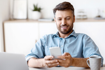 Poster - Happy Millennial Businessman Relaxing With Smartphone At Workplace In Office, Leaning Back In Chair, Texting With Friends Or Browsing Internet, Having Break In Work, Sitting At Desk With Laptop