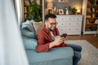 © Migma_Agency - Smiling man using smartphone and drinking coffee on sofa at home