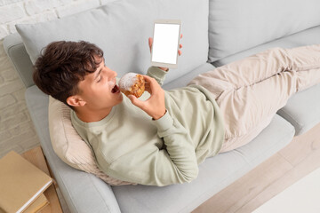 Wall Mural - young man with tablet computer eating tasty muffin on sofa at home