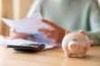 © Nanci - Woman managing home finances, calculating expenses and savings using calculator and reviewing bills, with piggy bank representing financial security