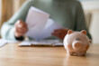 © Nanci - Senior woman reviewing domestic bills at home, sitting at a table with a piggy bank nearby, highlighting the importance of savings, financial planning, and managing retirement expenses