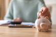 © Nanci - Senior woman managing finances by putting money in a piggy bank, calculating expenses, and planning future savings while checking documents at her desk