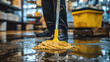 © Kanomaoi - A yellow mop cleans a wet, reflective floor, with a person and cleaning bucket visible in the blurred background.