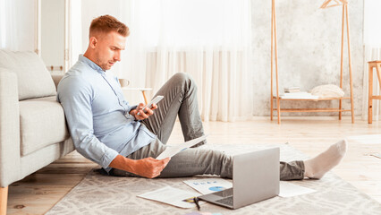 Poster - Young freelancer working from home, sitting on floor with pc, using smart phone and holding documents