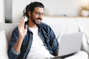 Poster - Online Communication. Positive Eastern Guy Making Video Call With Laptop At Home, Waving Hand At Web Camera, Young Arab Man Greeting Somebody While Sitting On Couch In Living Room, Free Space