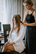 © Raivo - Hairdresser styling long straight hair of a young woman in a white robe during preparation for a special event at home..