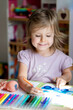 © 8th - Happy child girl paints a drawing in an album and smiles at home or in kindergarten at the table. Creative little artist at work. A charming girl is drawing with felt-tip pens at the table.