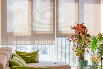 Poster - Automatic roller shades with screen material cover large windows, filtering sunlight into a living space. Motorized sunscreen blinds beige color with remote control panel on a sofa. Cityscape outside.