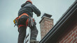 © dK - Chimney technician in safety gear on ladder inspecting residential brick chimney under cloudy sky