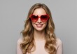 © Laki - Portrait of a smiling woman with blonde hair wearing red heart shaped sunglasses on a grey background