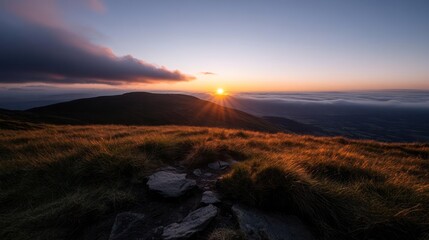 Fototapete - Golden sunrise over a grassy mountain top. The sun's rays burst from the horizon, casting a warm glow on the landscape with rocks in the foreground and hazy mountains in the distance. A dark cloud