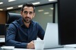 © Alex & M Studio - Smiling Middle Eastern businessman in a blue shirt working on a laptop in a modern office with large windows