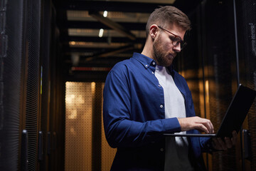 Poster - Man wearing glasses and blue shirt working on laptop in data center, surrounded by servers and technology equipment, concentrating on screen