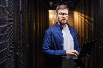 Poster - Portrait of man with glasses standing in server room holding laptop while managing servers. Wearing a blue shirt and analyzing server performance