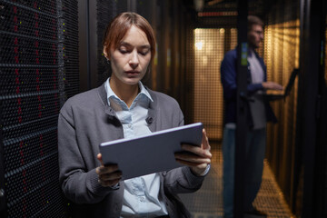 Poster - Woman working on tablet in data center with man in background using laptop. Focused on her tasks, highlighting modern technological environment and concentration on responsibilities