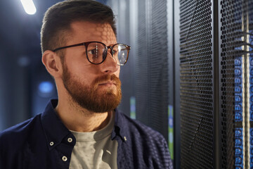 Poster - Bearded man standing in a data center, working with servers and equipment, wearing glasses and looking focused. Technology and servers visible in background