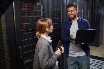 Poster - Image showing two IT professionals conversing in modern data center. Man holding laptop and woman holding tablet while standing in front of server racks