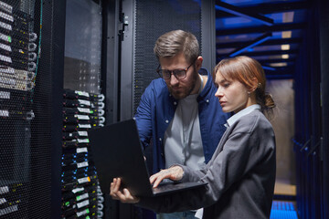 Poster - Two IT professionals are examining server connections in a data center, focusing on laptop screen while discussing necessary steps for troubleshooting issues