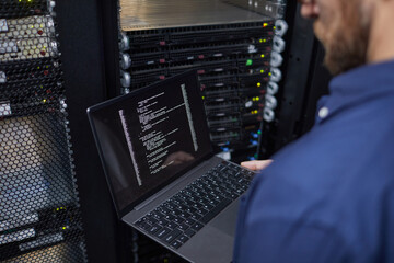 Poster - Technician standing in front of server rack holding laptop with terminal open displaying code. Focus on hands and laptop screen with server equipment in background