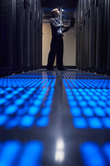 Poster - Technician wearing professional attire working on laptop inside server room, surrounded by advanced computer equipment and glowing blue lights on the floor creating a futuristic ambiance