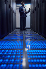 Poster - Man holding laptop in a data server room, surrounded by tall server racks, with blue illuminated floor panels creating a futuristic atmosphere