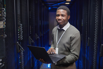 Poster - IT professional standing in server room, holding laptop with confident expression. Blue lighting highlights server racks in background, indicating technology environment