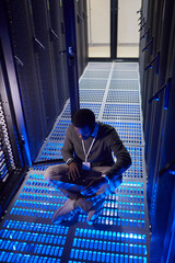 Poster - Engineer monitoring server performance while sitting on floor inside data center filled with blue neon lighting. Surrounded by towering server racks, focused on device