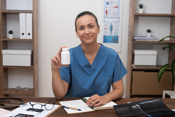 Poster - Smiling female doctor holds bottle with capsules sitting at desk in clinic woman in medical uniform recommends innovative medicines prescription for patient treatment