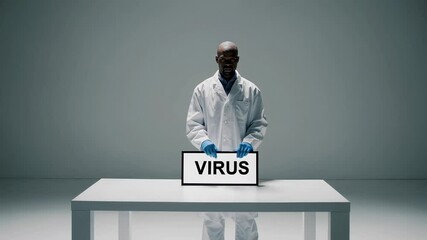 Poster - Serious virologist wearing lab coat and gloves shows a sign with the word virus written on it while standing behind a table on a gray background