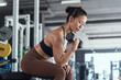 © 2B - Focused woman lifts dumbbell while sitting on bench in modern gym during daytime workout session