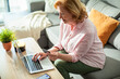 © Marko Geber - Senior woman sitting on couch online shopping with laptop at home