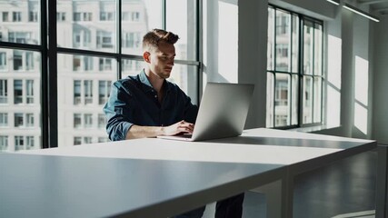 Aufkleber - Professional businessman focusing intently while working on laptop, sitting at sleek desk with bright sunlight streaming through expansive office windows