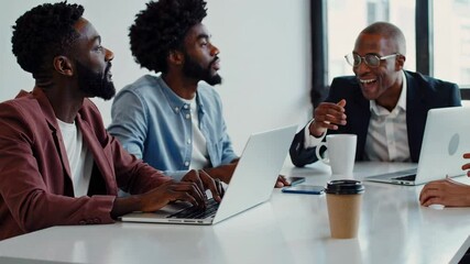 Fototapete - Three businessmen are sitting at a table, discussing a project, with two of them looking at their laptops and the other one gesturing while speaking