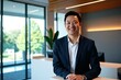 © Alex & M Studio - happy Asian businessman in a smart dark suit and white shirt stands in a modern, well-lit office with large windows showing a green outdoor scene, smiling confidently at the camera