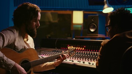 Leinwandbilder - Songwriter composing a song with sound engineer in professional studio, recording session with his electro acoustic guitar in control room. Musician producing tracks with mixing console. Camera B.