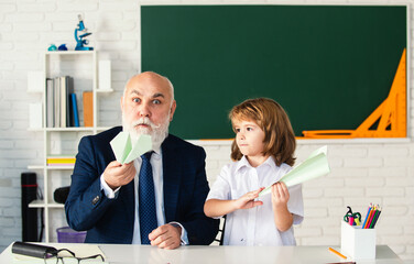 Fototapete - Happy kid in school break. Senior teacher with pupil with paper plane in school classroom. Education, elementary school. School break. Teacher with pupil with airplanes. Education kids concept.