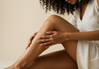 © kristina - Young African American woman in white dress gently touching her smooth legs while sitting in minimal studio setting, representing skincare, softness, body care and feminine beauty in natural light