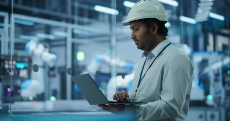 Fototapete - Indian Male Engineer in a Hard Hat Using a Laptop Computer to Monitor Robotic Arms in an Advanced Industrial Factory. South Asian Specialist Overlooking Modern Automated Electronics Manufacturing