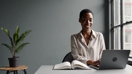 Aufkleber - Office worker smiles while typing on her laptop with a notebook open in front of her, near a window with natural light and a plant on a small table