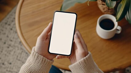 Poster - Woman wearing beige sweater holding smartphone with white screen near coffee cup and green plant, working on wooden table in cozy indoor environment