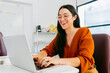 © juanpablo - In a workspace, a young Latina woman happily types on a laptop computer