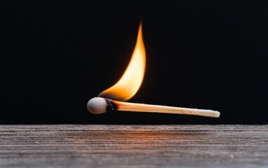 Poster - Lit matchstick hovering above a rustic wooden surface against a black background. The flame is bright and dynamic, casting a warm glow