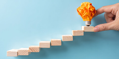 Poster - Orange paper lightbulb on wooden steps, hand placing it, against light-blue background, symbolizing idea development and progress, achievement