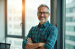 © Vadym - Mature businessman with crossed arms smiles at camera. Man in glasses stands by office window. Happy pro in buttoned shirt, grey hair, confident expression.