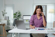 © crizzystudio - Smiling businesswoman sitting at office desk, using calculator and talking on smartphone while analyzing financial statistics displayed on documents and laptop