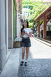 © crizzystudio - Happy young asian woman walking down a bustling city street, using her smartphone to connect with friends while enjoying a sunny day, radiating joy and independence