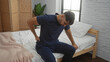 © Krakenimages.com - Young man sitting on bed in home bedroom experiencing back pain with a thoughtful expression, framed by cozy interior elements including pillows and soft lighting.