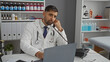 © Krakenimages.com - Young handsome male doctor in a clinic room talks on the phone while working with a laptop, surrounded by medical supplies, conveying a professional healthcare setting.