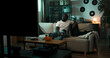 © ABCreative - Man sits on the couch holding a bowl of chips, flipping through sports channels. He relaxes in the dim room lit by a lantern outside the window, searching for a game to watch.