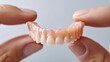 © NovaVision - Hands delicately holding a set of upper dentures against a clean, light background in close-up.